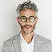 Smiling middle-aged man with curly gray hair, glasses, and beard wearing a light gray blazer.
