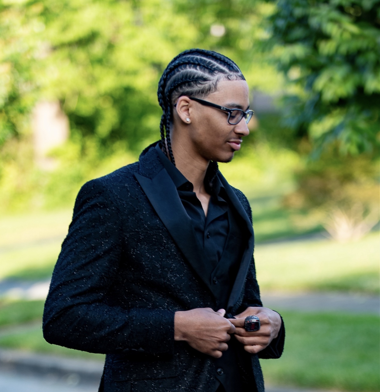 Young man with braided hair wearing glasses and a black textured blazer buttoning his jacket outdoors.