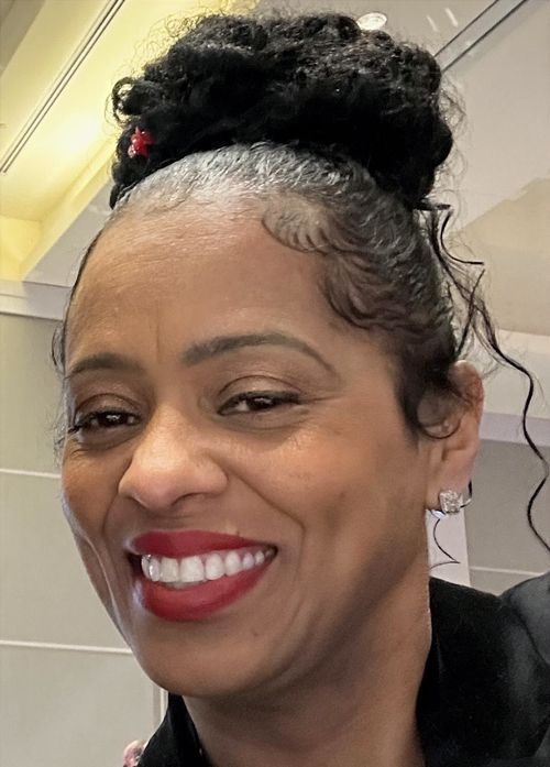 Close-up of a smiling woman with dark curly hair styled in a bun, wearing red lipstick and diamond earrings.