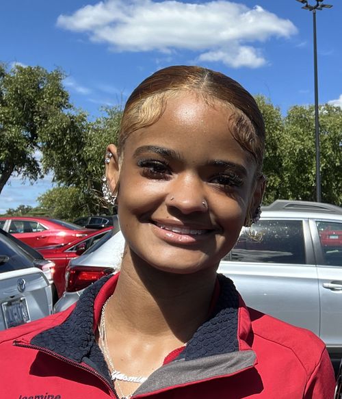 Smiling woman with styled baby hairs, nose piercings, and multiple ear piercings wearing a red jacket and a necklace that reads 'Jasmine' in a parking lot under blue sky with clouds.