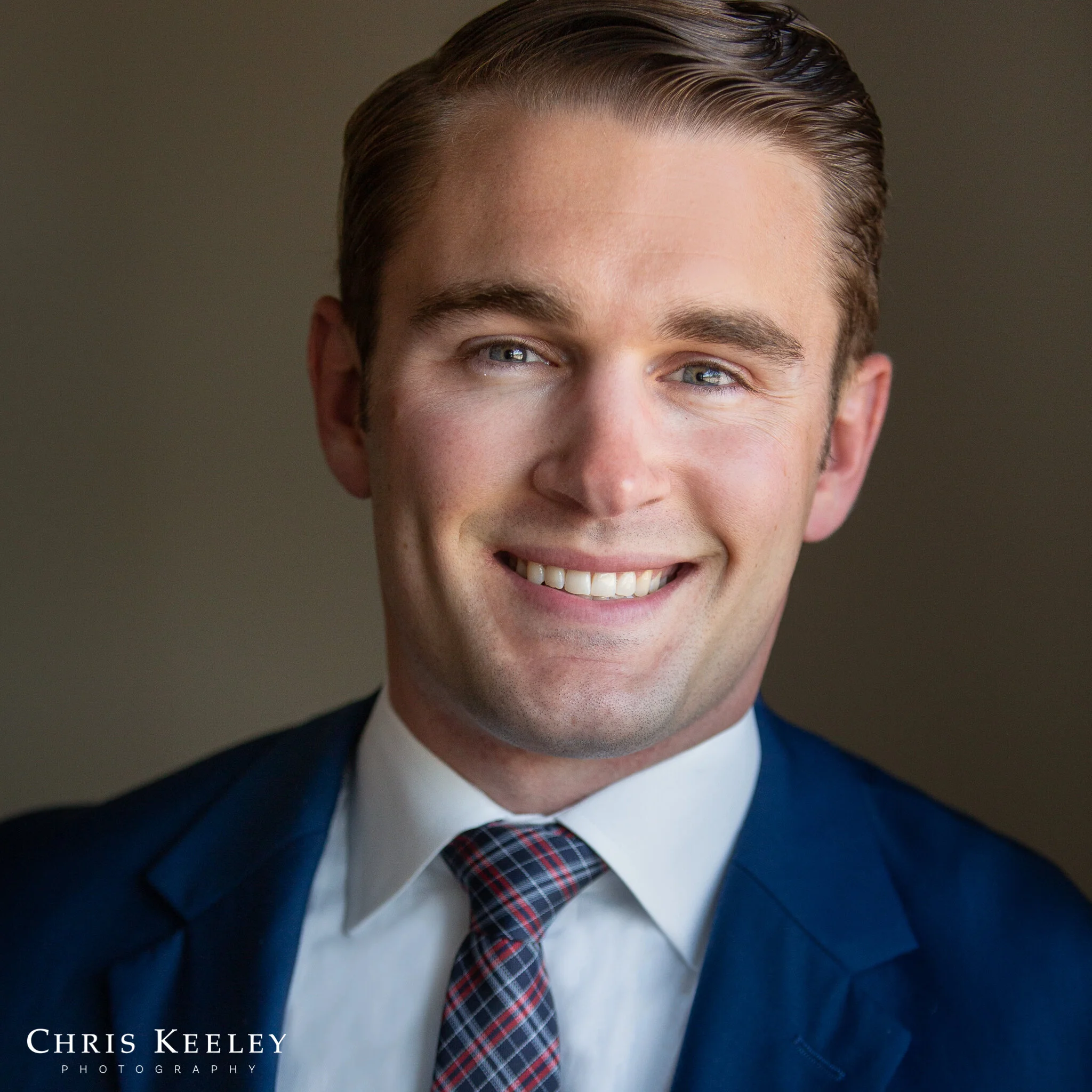 Smiling man with slicked-back brown hair wearing a blue suit jacket, white shirt, and plaid tie against a plain background.