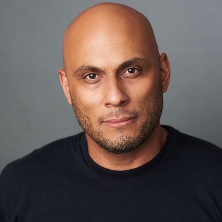 Close-up portrait of a bald man with a trimmed beard wearing a black shirt, against a gray background.