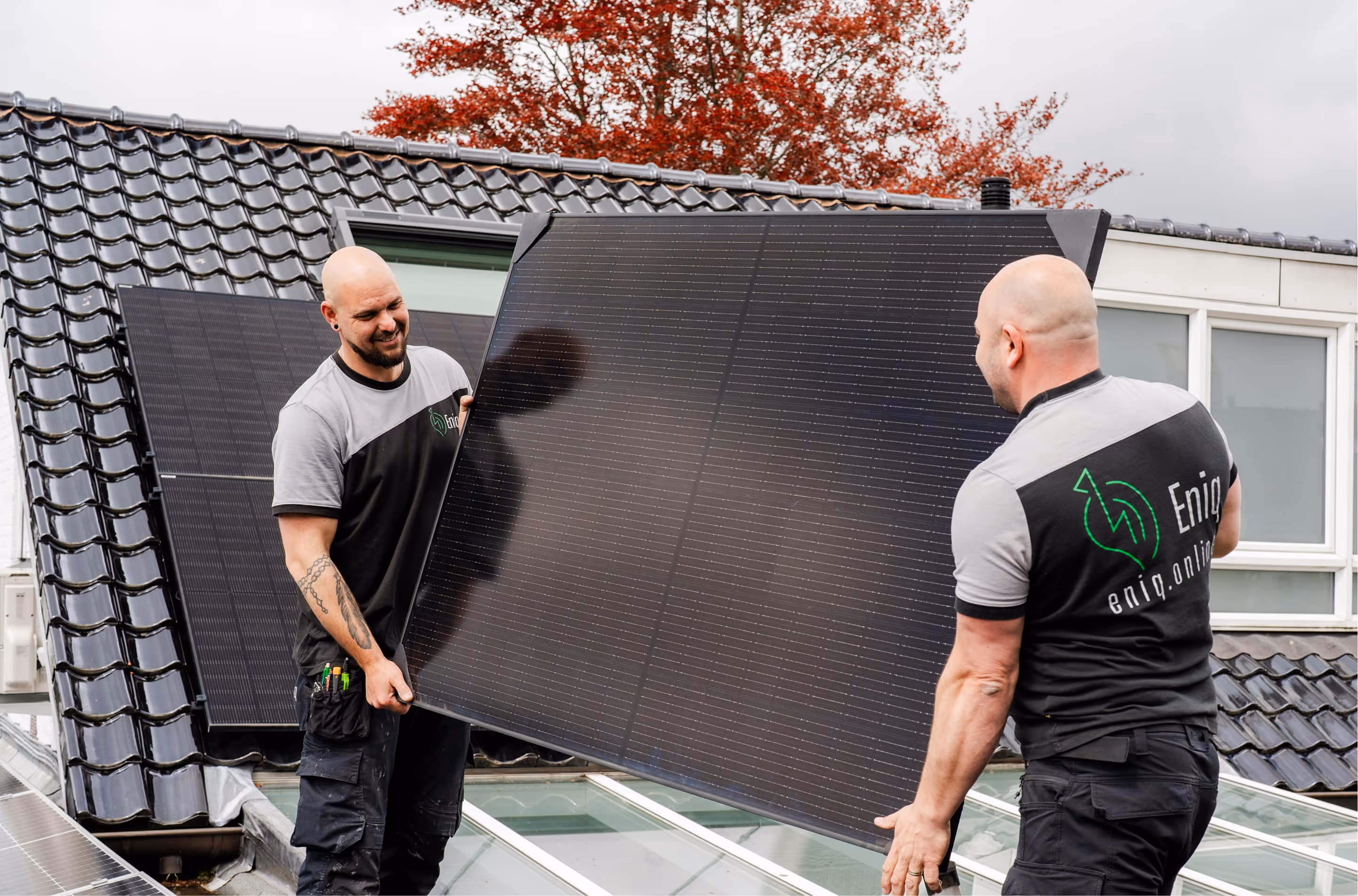 Twee mannen installeren een zonnepaneel op een dak met zwarte dakpannen en een boom met rode bladeren op de achtergrond.