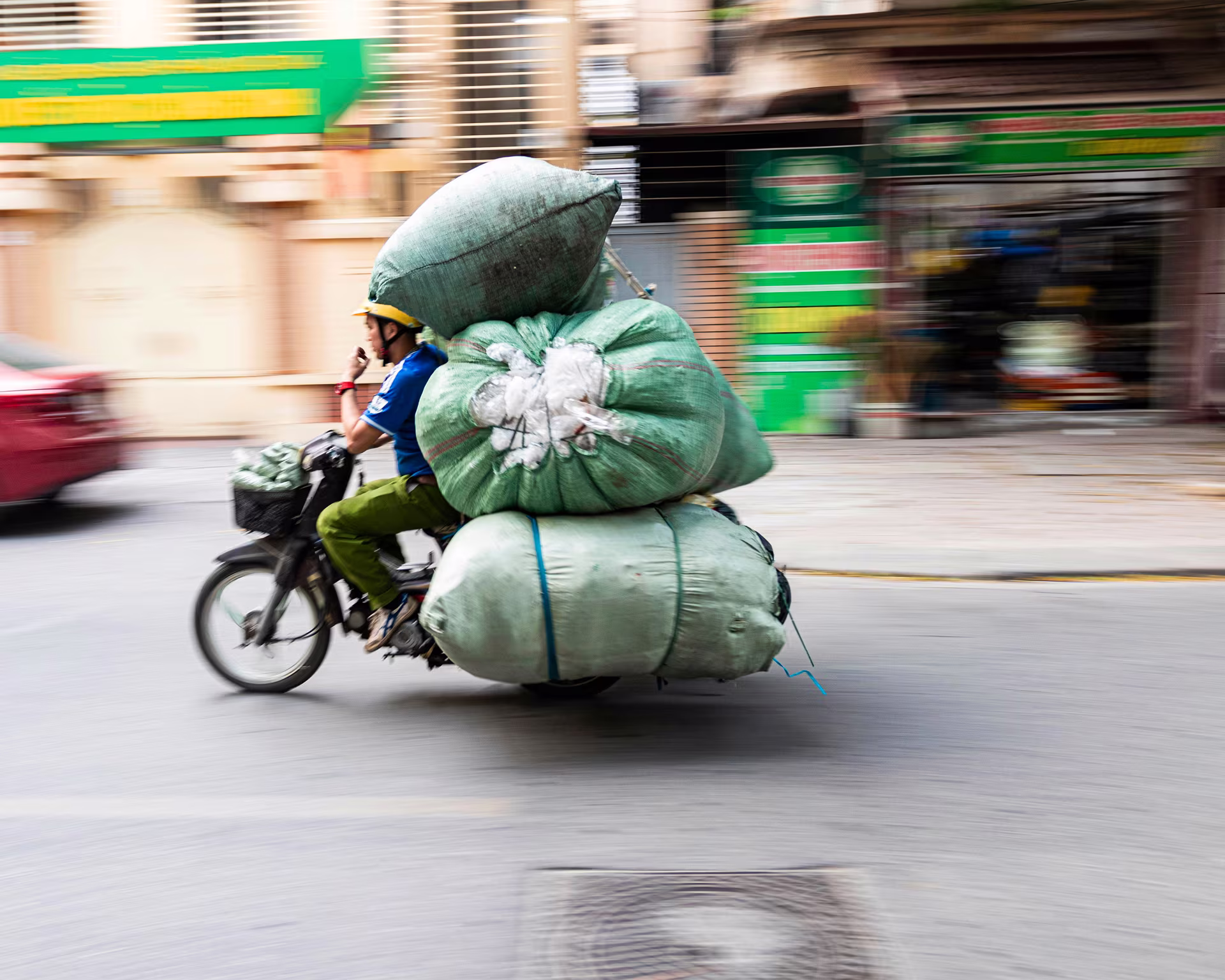 Man riding a motorbike heavily loaded with large green bags on a city street.
