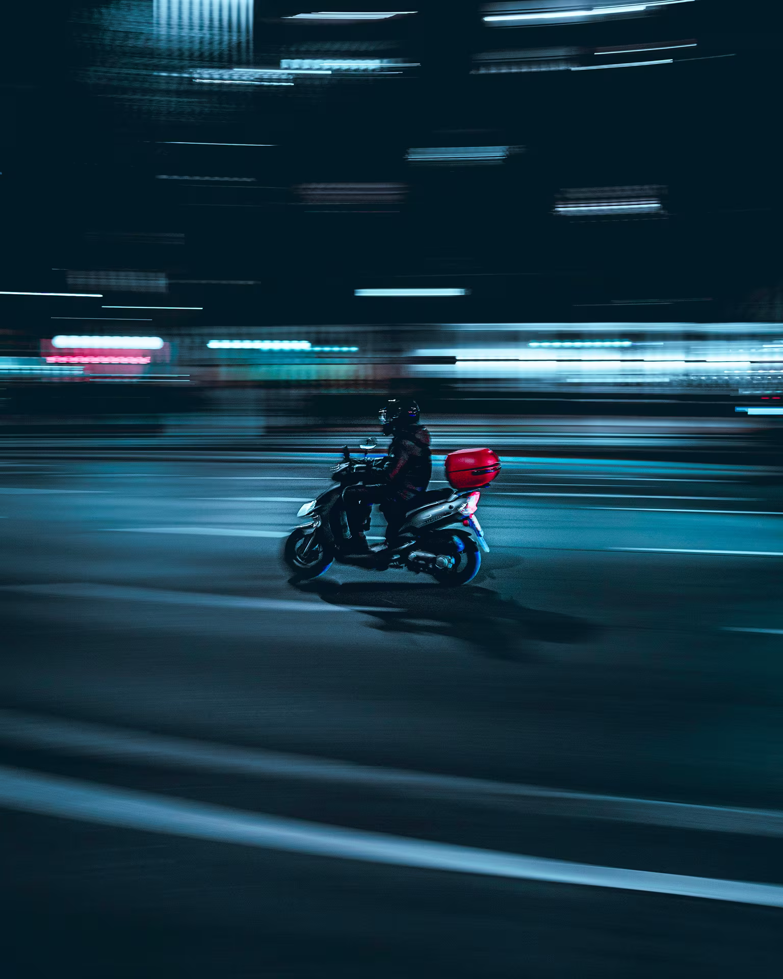 Person riding a scooter with a red storage box at night on a city street with blurred lights.