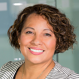 Smiling woman with curly short hair wearing a striped top and necklace against a blurred indoor background.