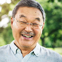 Smiling middle-aged Asian man with glasses wearing a light blue checkered shirt outdoors.
