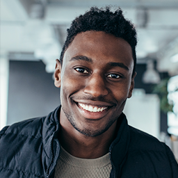 Smiling young man with short curly hair wearing a black jacket over a beige shirt in a modern indoor setting.