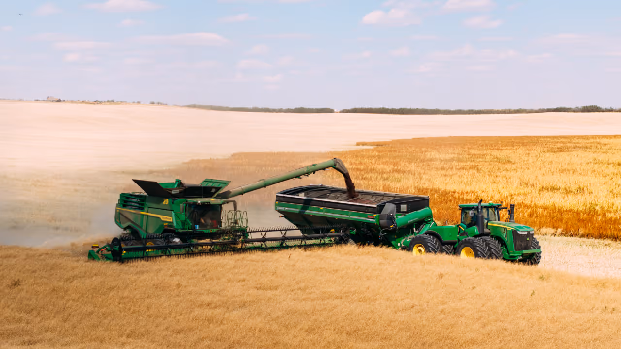 Green combine harvester unloading grain into a green tractor trailer in a golden wheat field under a partly cloudy sky.