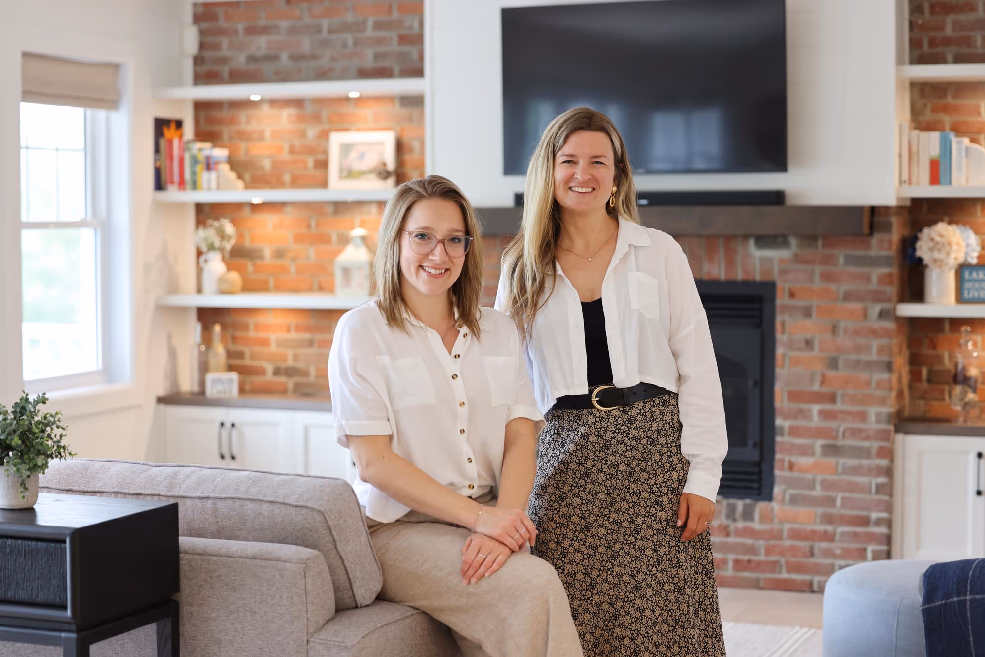 Deux femmes souriantes dans un salon moderne avec mur en briques, télévision, et étagères décorées.