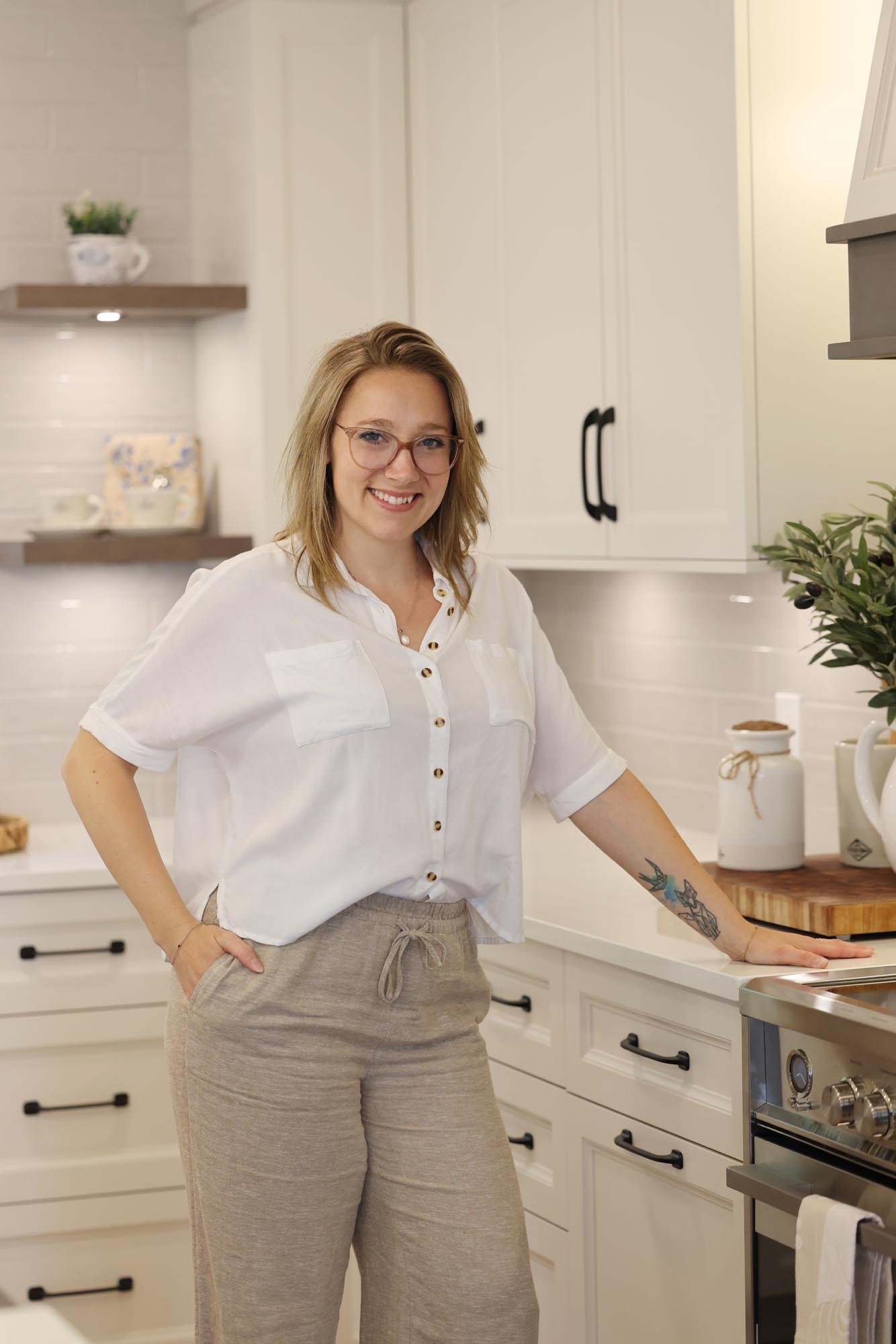 Femme souriante portant des lunettes et une chemise blanche, debout dans une cuisine moderne avec des armoires blanches.