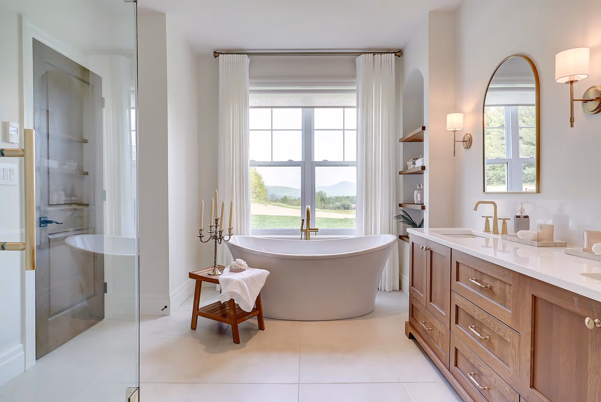 Salle de bain lumineuse avec une baignoire moderne devant une fenêtre, un meuble-lavabo en bois et un miroir ovale mural.