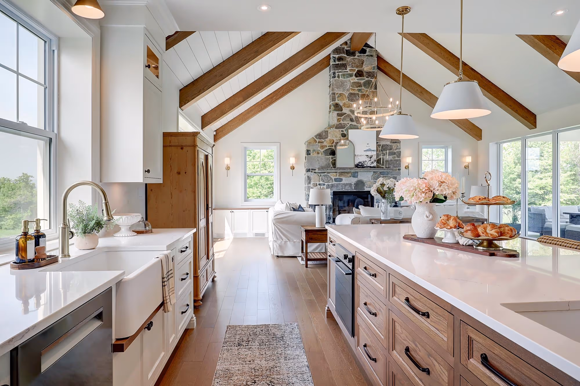 Cuisine lumineuse au style ferme moderne avec îlot en bois, poutres apparentes au plafond et cheminée en pierre dans le salon adjacent.