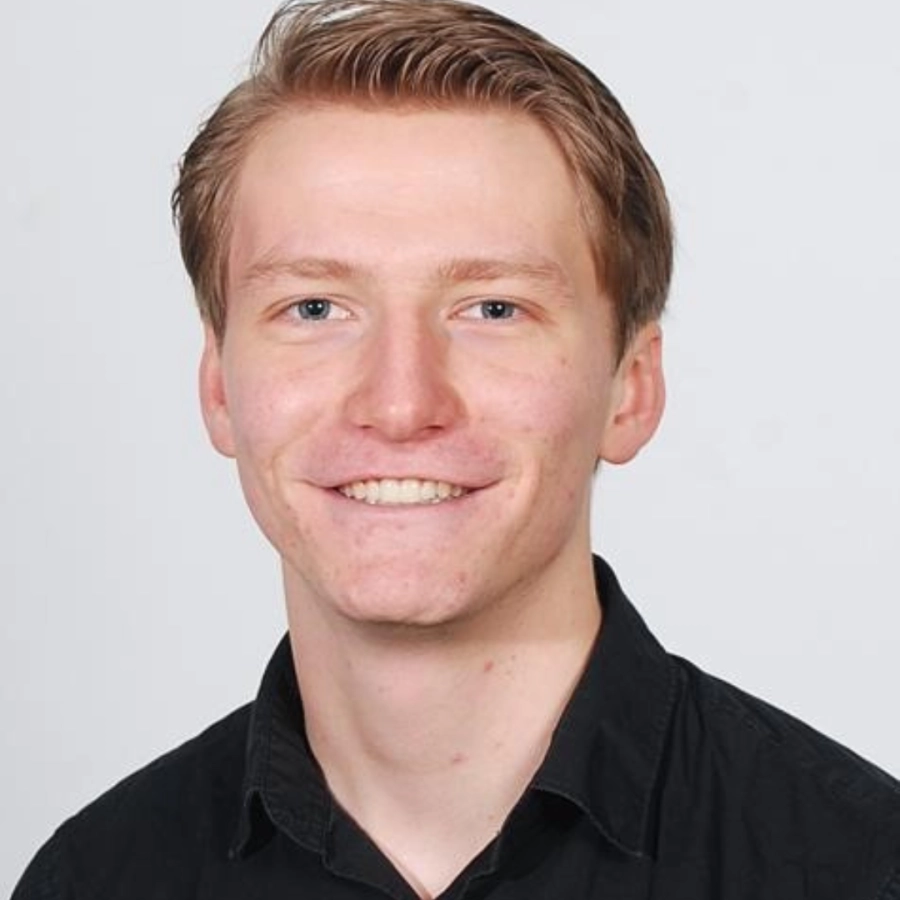 Portret van een glimlachende man met kort lichtbruin haar, gekleed in een zwart shirt tegen een lichte achtergrond.