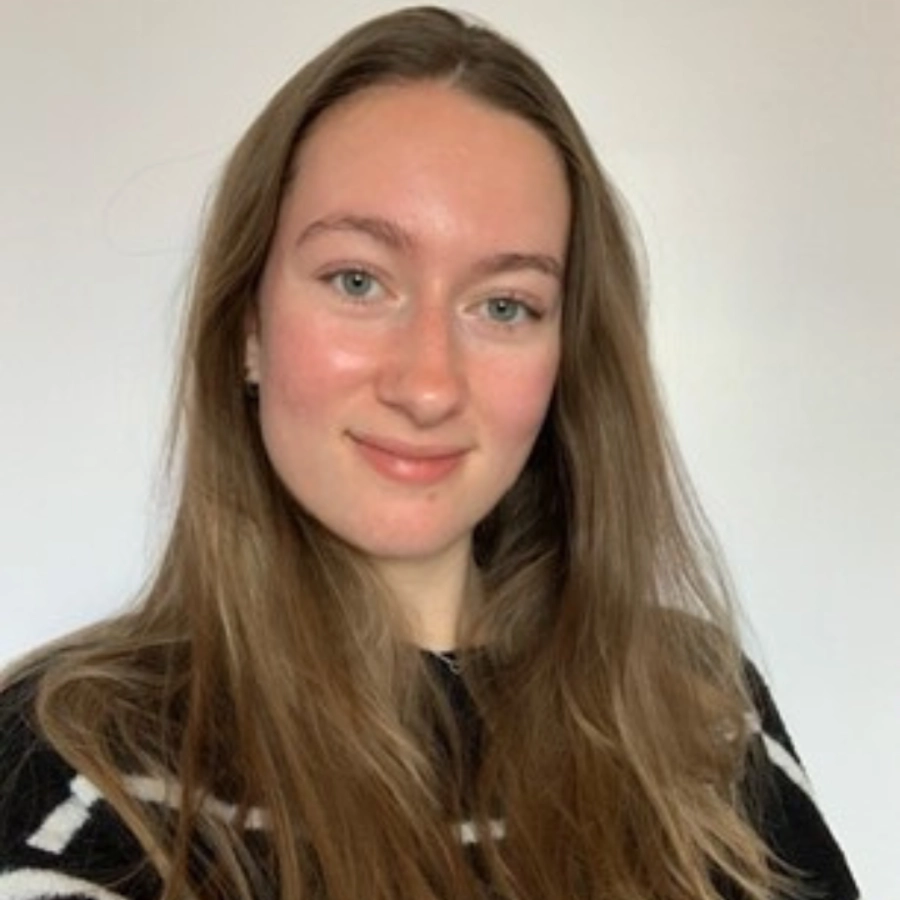 Portret van een vrouw met lang lichtbruin haar, met een lichte glimlach, gekleed in een donkere top tegen een effen lichte achtergrond.