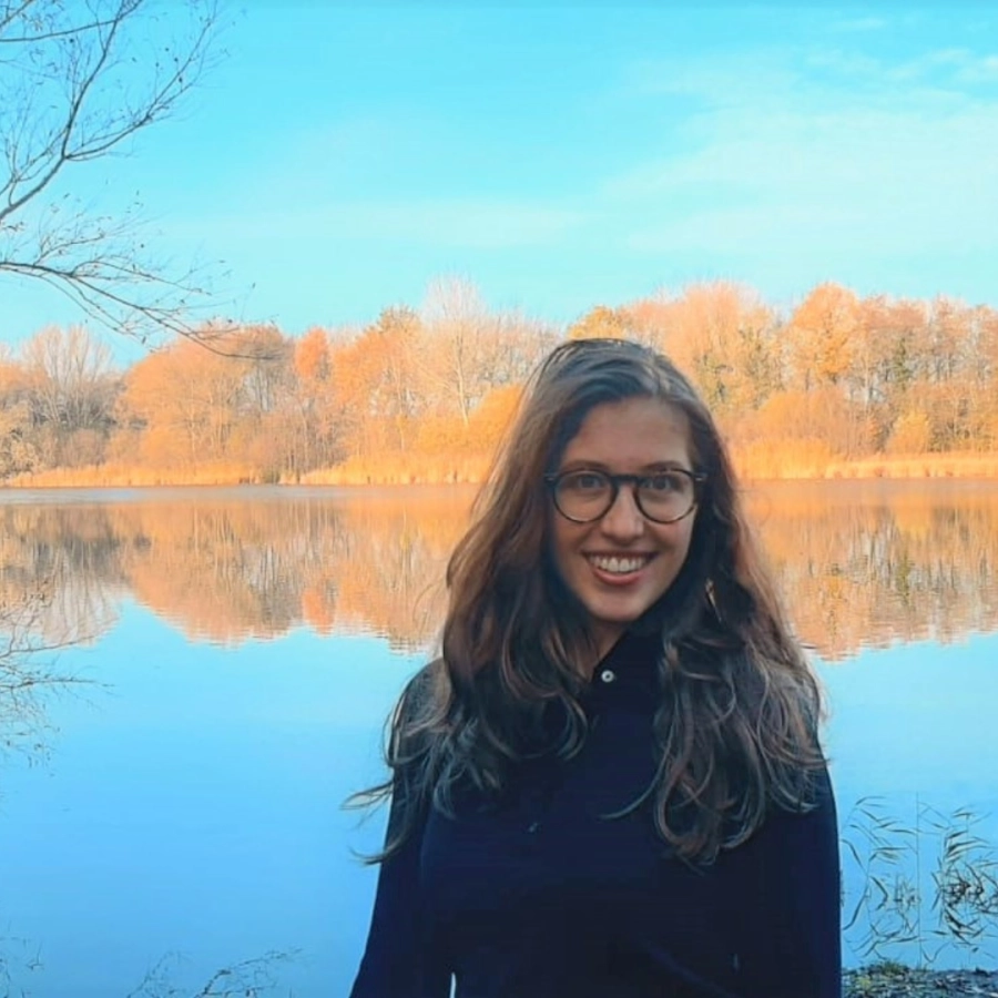 Een vrouw staat bij een rustig meer met herfstbomen die weerspiegeld worden in het water.