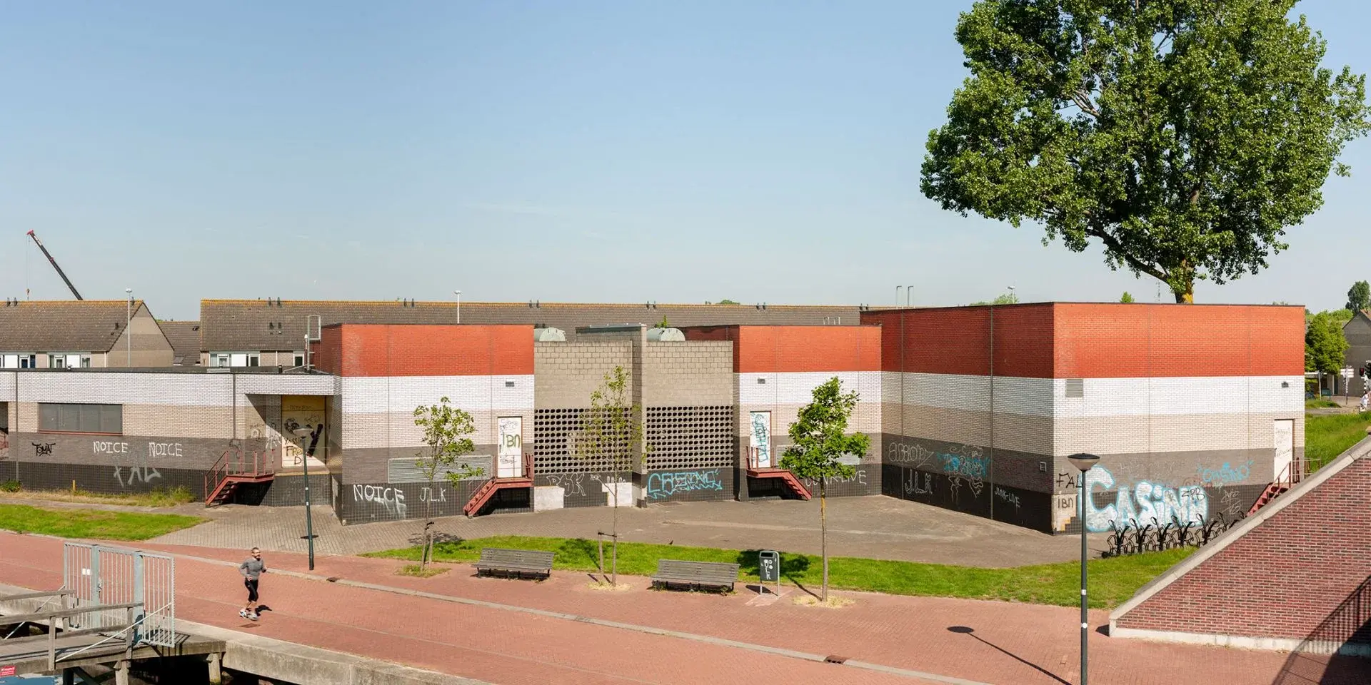 Straatbeeld van een laag bakstenen gebouw met horizontale banen in rood, wit en grijs, graffiti op de gevel, een fiets- en wandelpad ervoor, bankjes, jonge bomen en omliggende woonbebouwing.