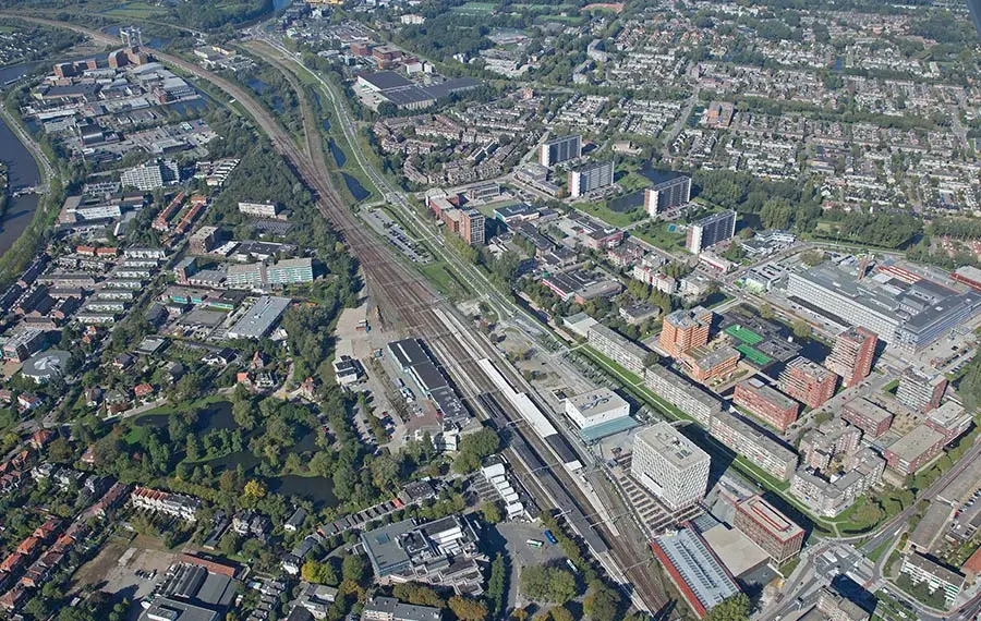 Luchtfoto van de stad Gouda met het centraal gelegen treinstation en meerdere spoorlijnen, omringd door appartementen, wegen, groenvoorzieningen en woonwijken.