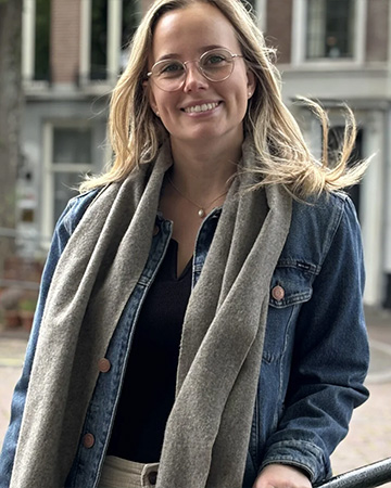 Portret van een glimlachende vrouw met een bril, een spijkerjack en een sjaal, staand buiten op een klinkerstraat met woongebouwen op de achtergrond.