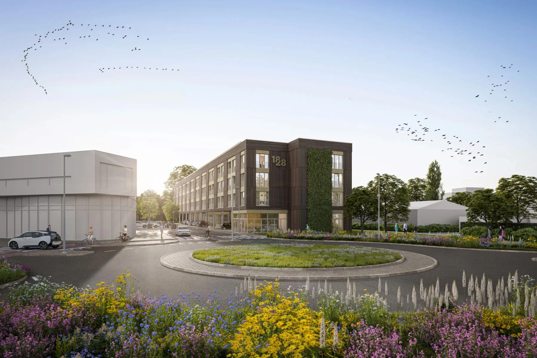 Straatbeeld van een groen ingerichte rotonde met kleurrijke wilde bloemen, fietsers en voetgangers, en een modern woongebouw op de achtergrond, gelegen in een groene stedelijke omgeving met vogels in de lucht.