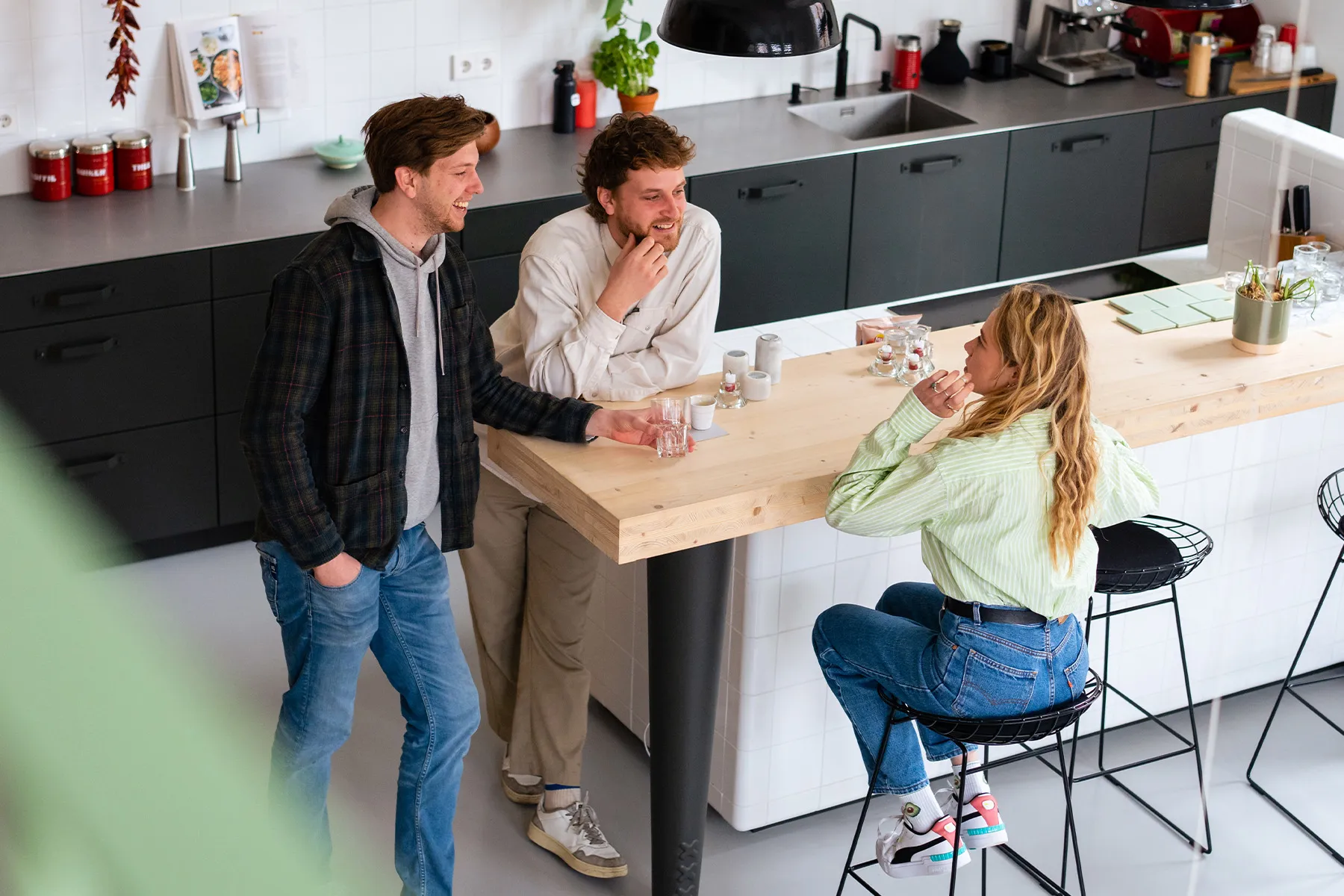 Drie jonge mensen die met elkaar praten en lachen rond een kookeiland, één persoon zit op een barkruk terwijl de anderen staan met drankjes in een lichte, moderne keuken.