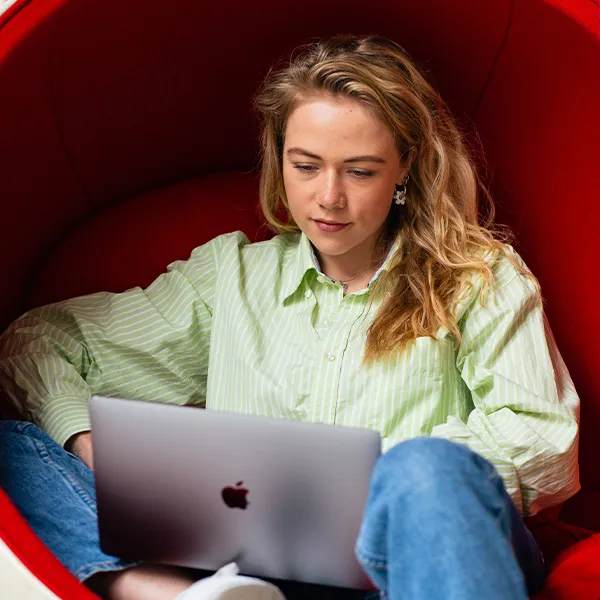 Jonge vrouw die in een rode loungestoel zit en op een laptop werkt, gekleed in een lichtgroen gestreept overhemd en een spijkerbroek.