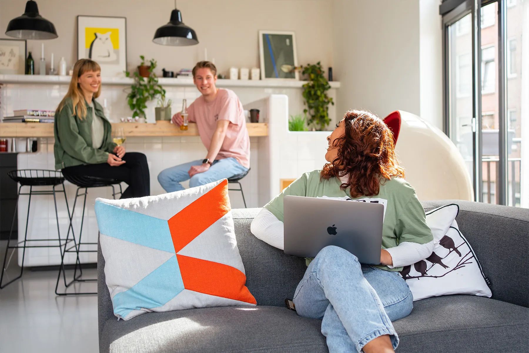 Groep jonge mensen binnen die met elkaar in interactie zijn: een vrouw zit op de bank met een laptop en praat met twee anderen bij het keukenblad op de achtergrond.