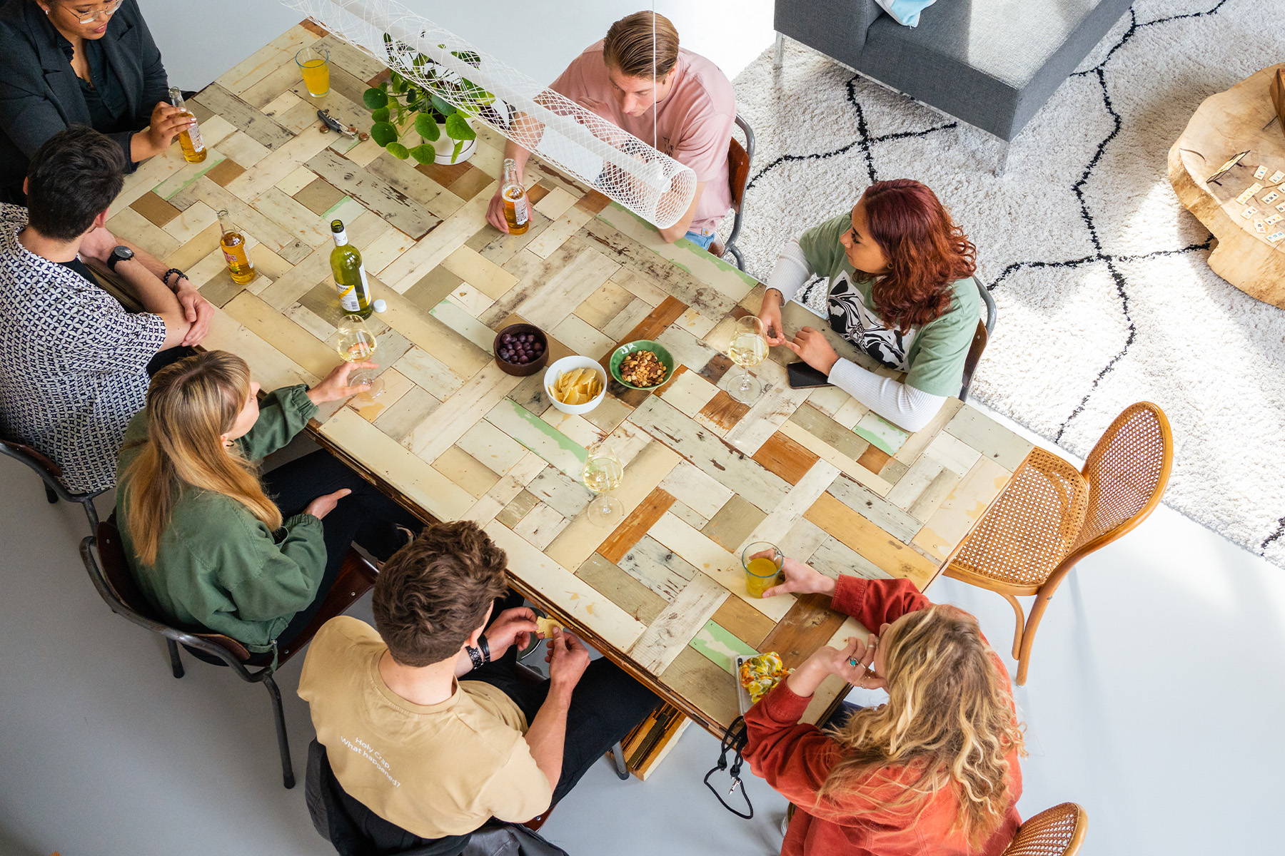 Bovenaanzicht van een groep bewoners die samen aan een grote houten tafel zitten, praten en hapjes en drankjes delen in een lichte gemeenschappelijke ruimte.