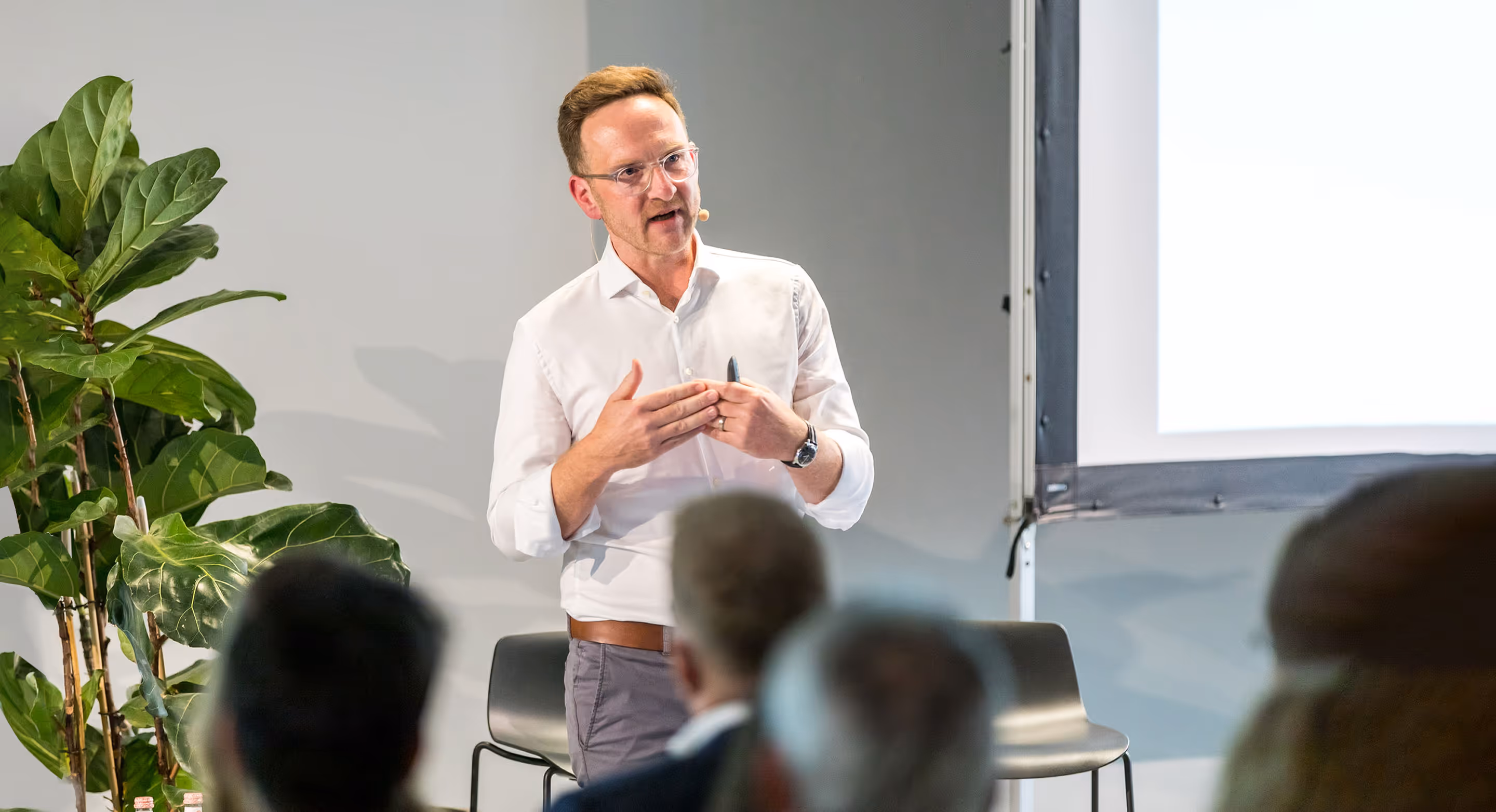 Ralph Steffens spricht vor einem Publikum mit Mikrofon, steht vor einer Leinwand und einem grünen Pflanzenarrangement.