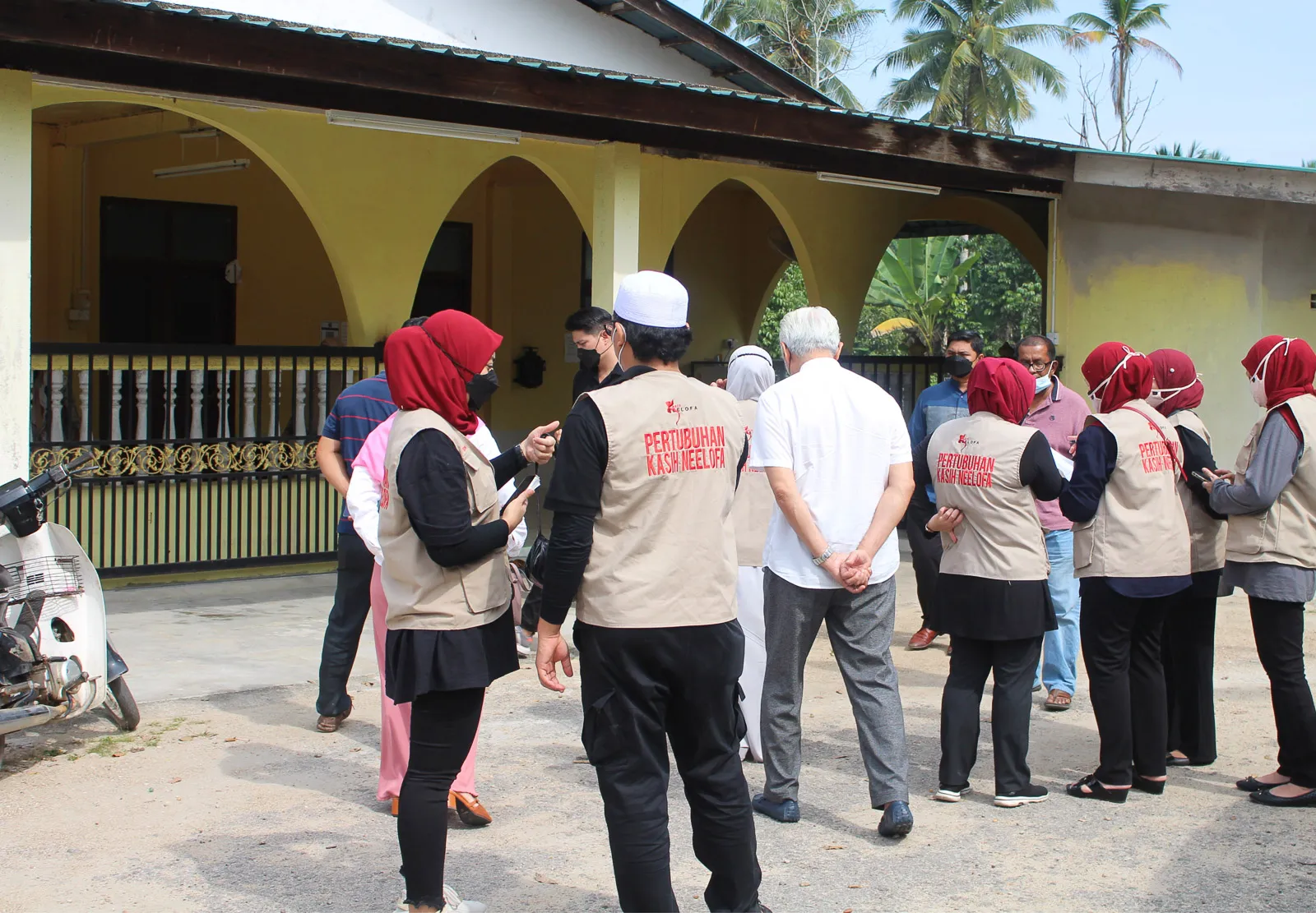 Group of people wearing Pertubuhan Kasih Neelofa vests gathered outside a building with yellow walls and arches.