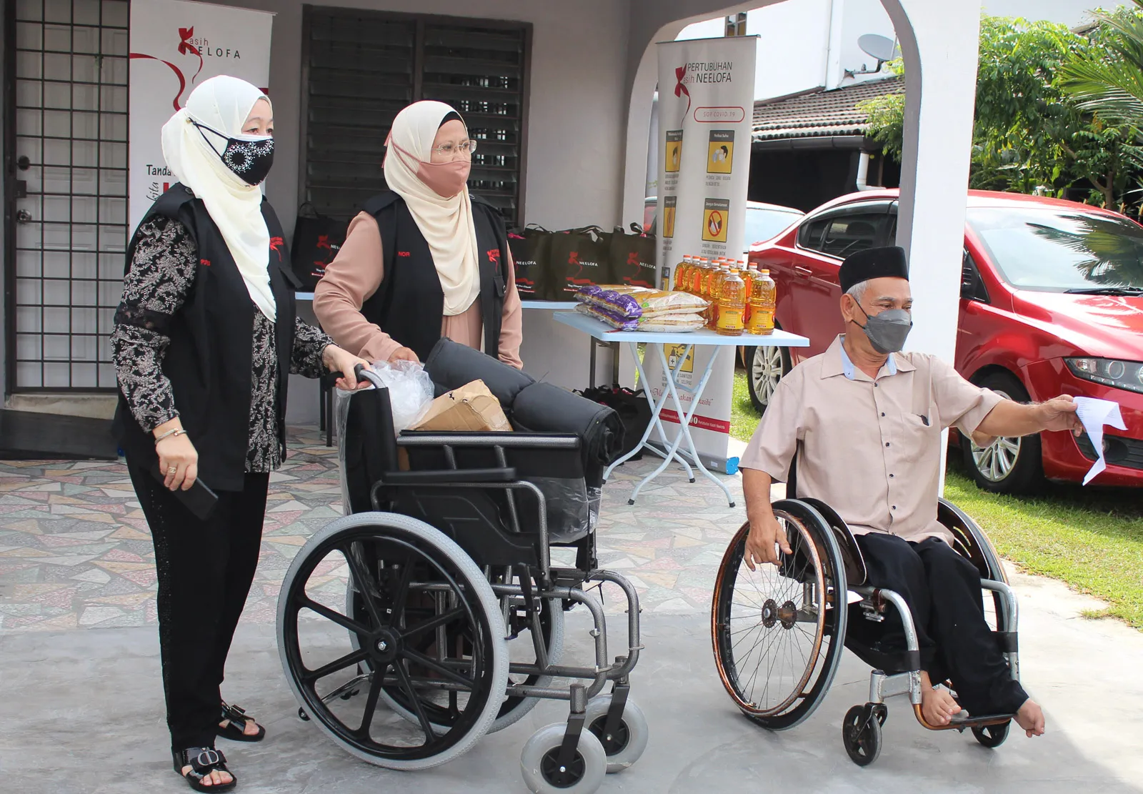 Two women wearing hijabs and face masks assist a man in a wheelchair outdoors near a table with groceries and supplies.
