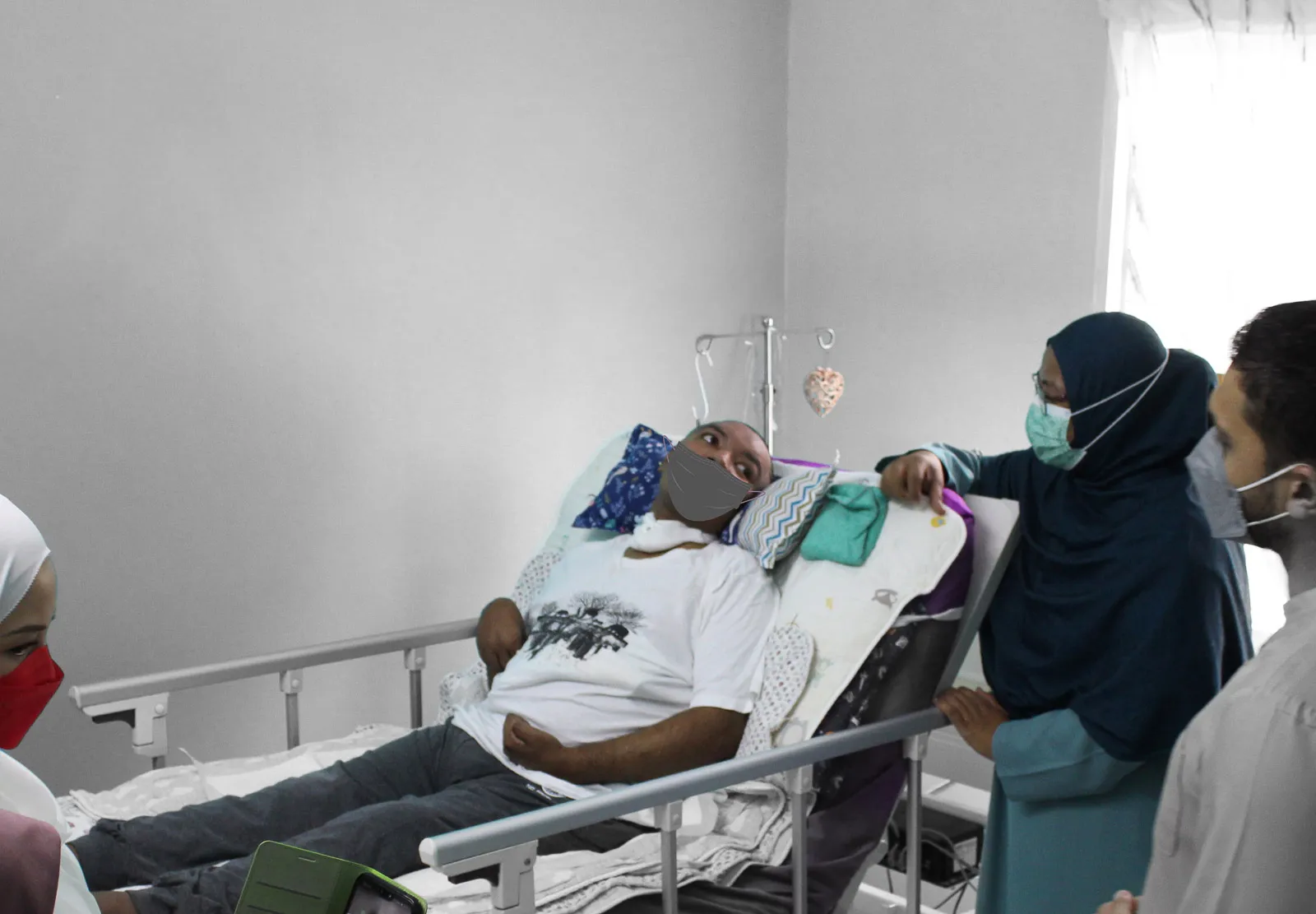 Man wearing a face mask lying in a hospital bed, accompanied by three visitors also wearing face masks.