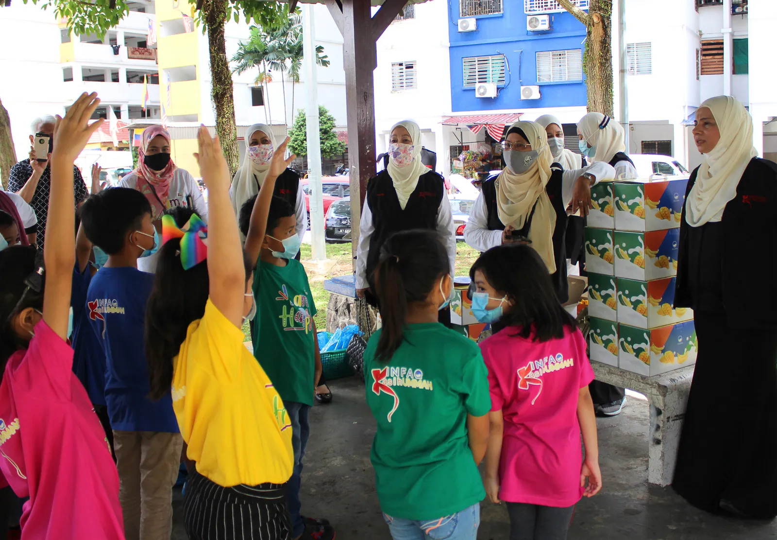 Group of children wearing colorful shirts and face masks raising their hands in front of a group of women in headscarves and face masks under a shelter with boxes stacked on a bench.
