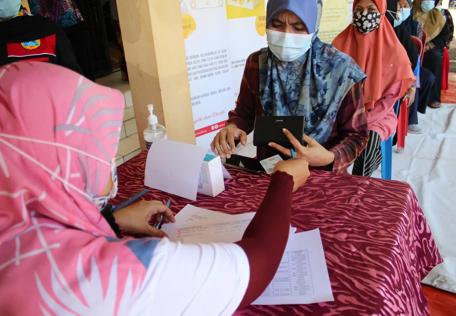Women wearing face masks waiting in line at a registration table where a woman is checking an ID card and documents.