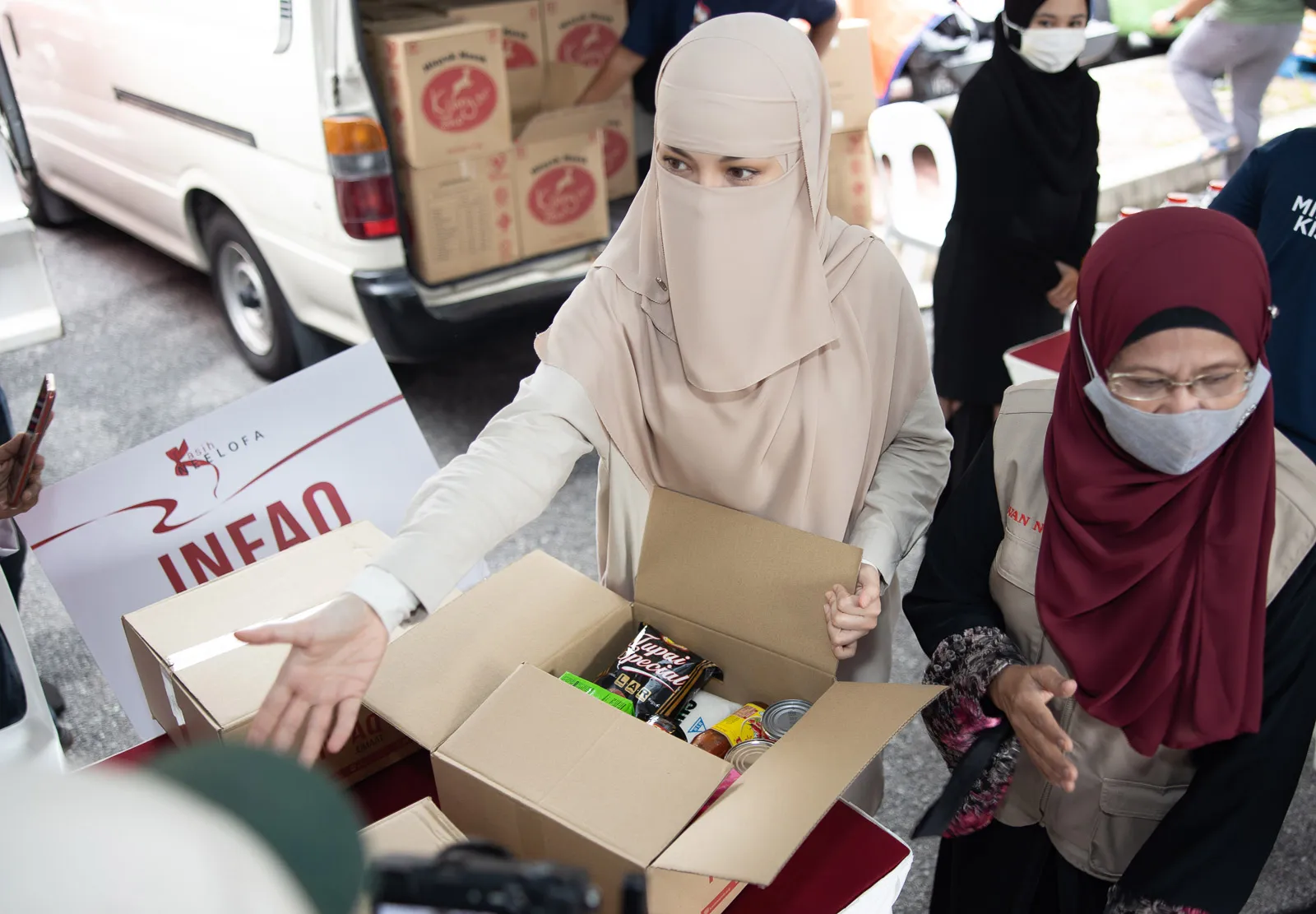 Woman wearing a beige niqab distributing a box of food supplies during a community aid event.
