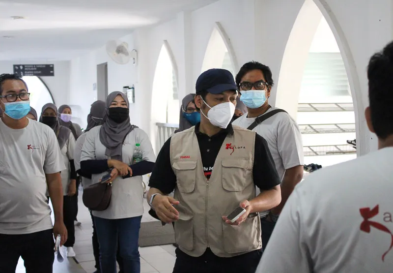 Group of masked volunteers walking in a building corridor, one man in front wearing a beige vest and holding a phone.