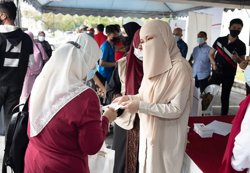 Women wearing hijabs and face masks interacting and exchanging cards at an outdoor event with people in the background.