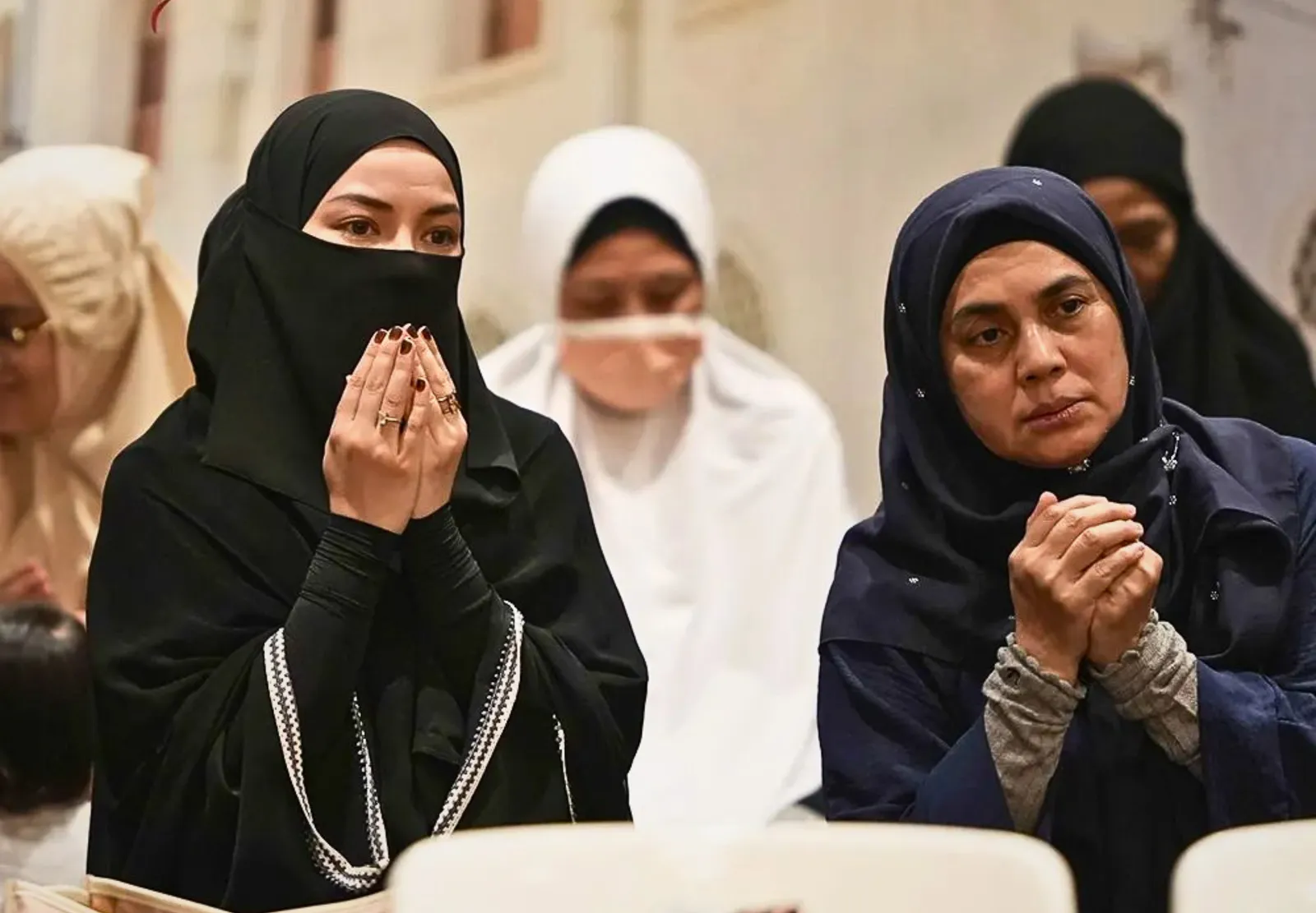 Two Muslim women wearing hijabs praying with hands raised indoors, with others praying in the blurred background.