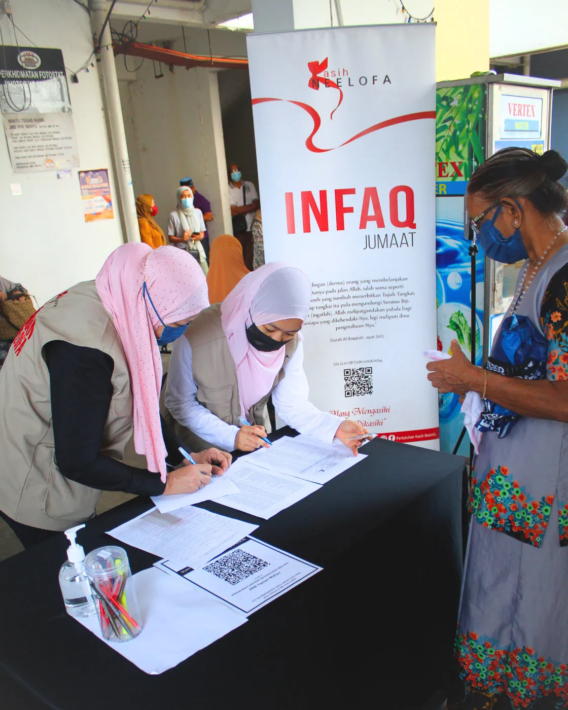 Two women in hijabs and face masks signing documents at a donation table with a banner reading 'INFAQ JUMAAT' and a woman standing opposite them.