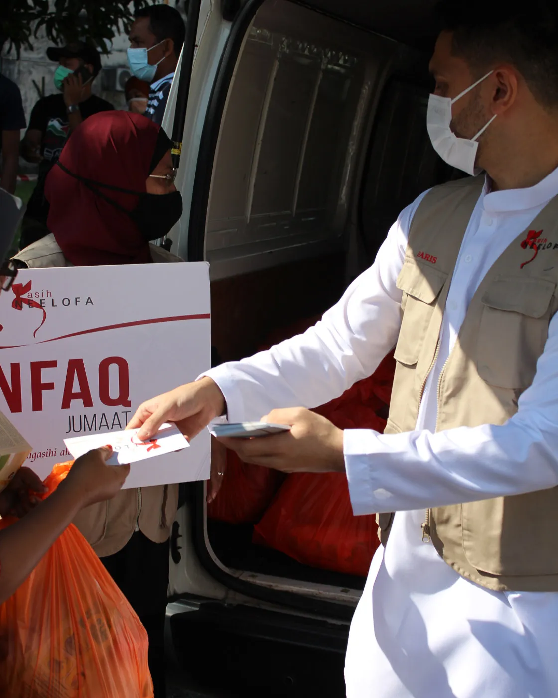 Man in white outfit and face mask handing out donation envelopes near a van with red donation bags and a woman in a headscarf holding a large sign.