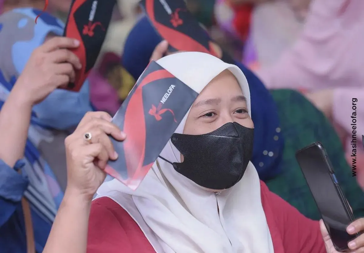 Woman wearing a white hijab and black face mask holding up a card with Neelofa logo and a smartphone.