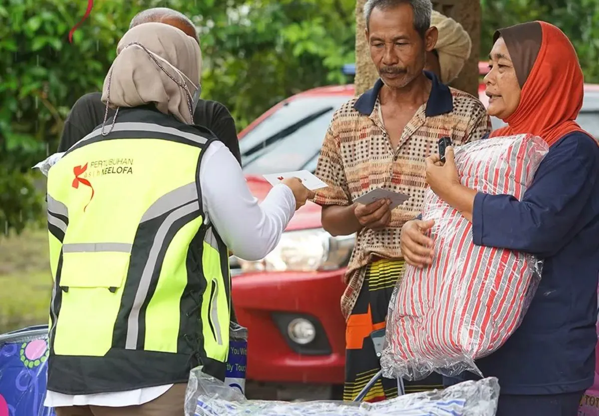 Person in a reflective yellow vest handing out cards to a man and woman holding a striped plastic package outdoors.