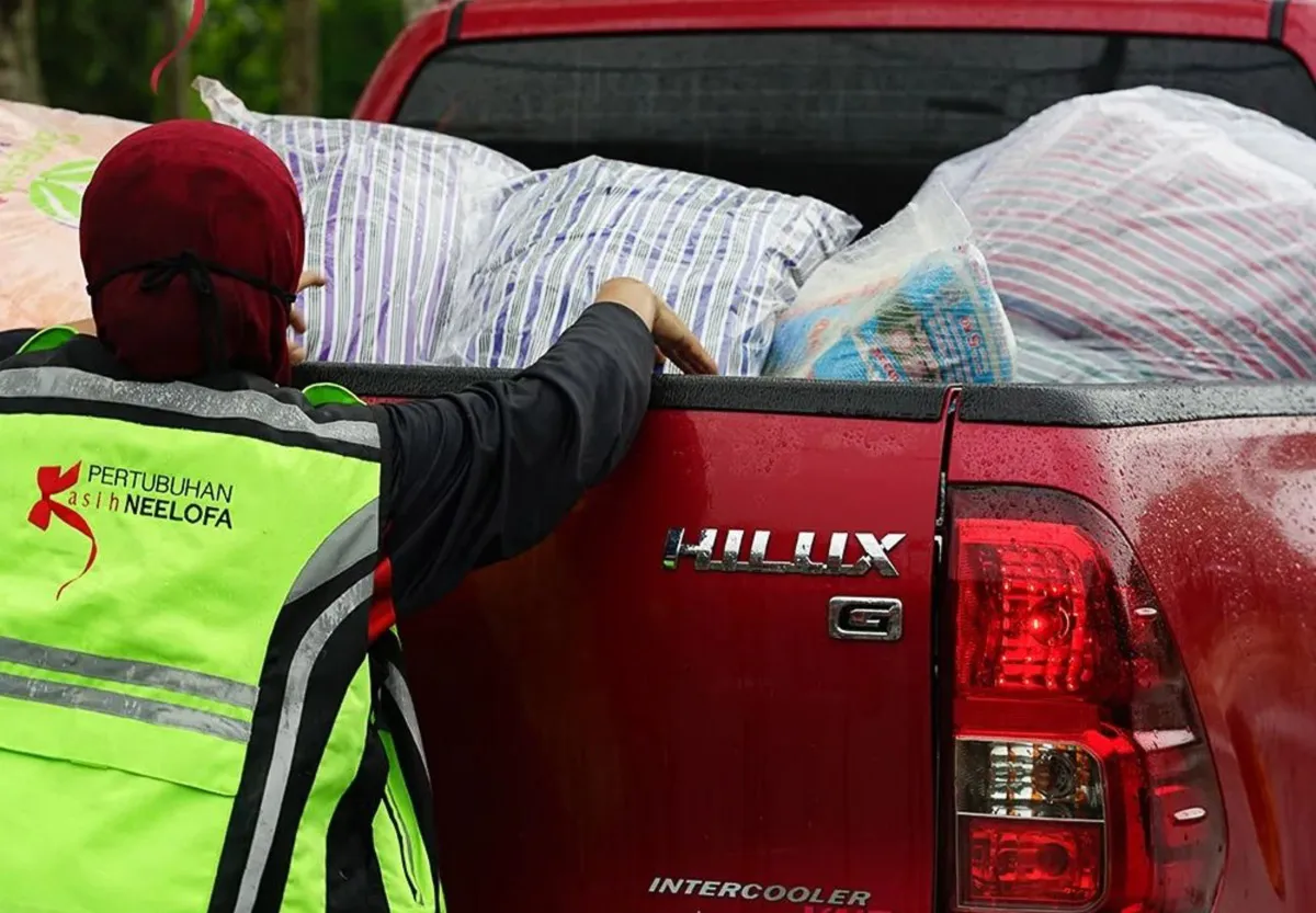 Person wearing a Pertubuhan Kasih Neelofa safety vest loading bags into the back of a red Toyota Hilux pickup truck on a rainy day.
