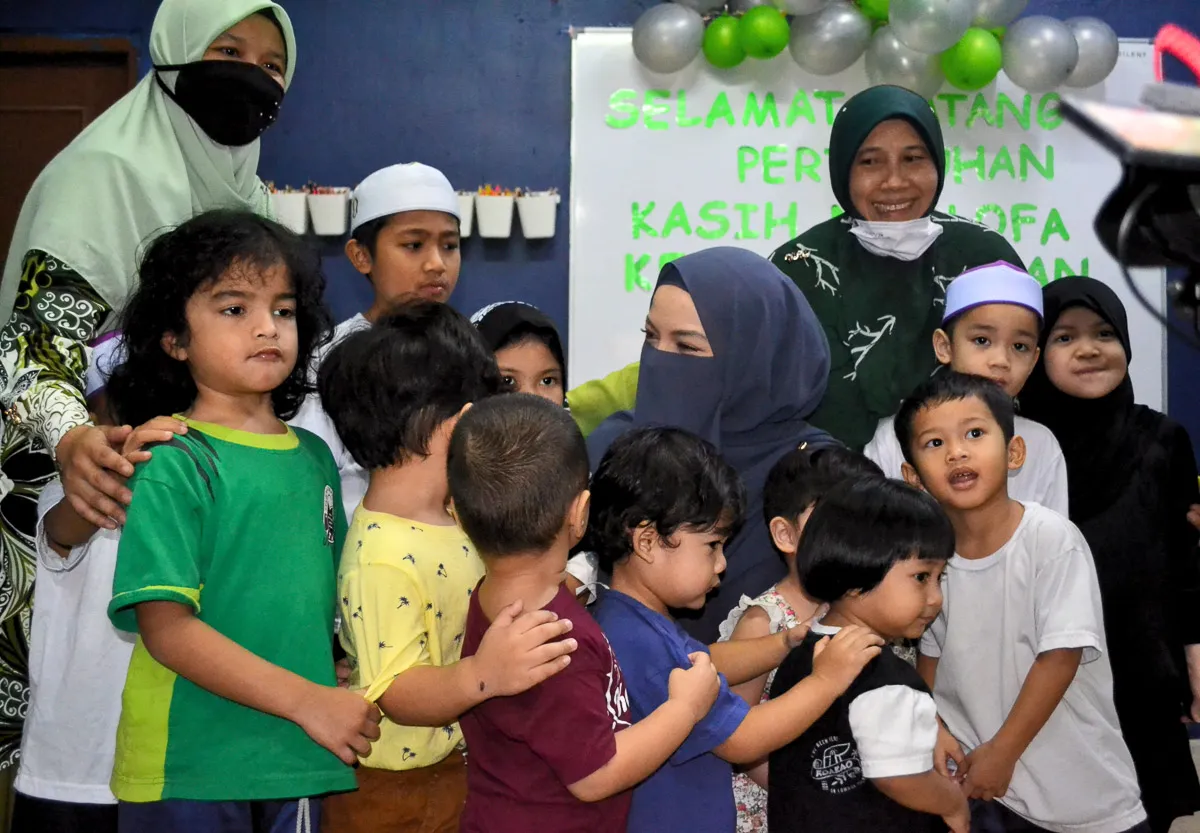 Group of children and two women in hijabs gathered indoors with green and silver balloons and a welcome sign in Malay behind them.
