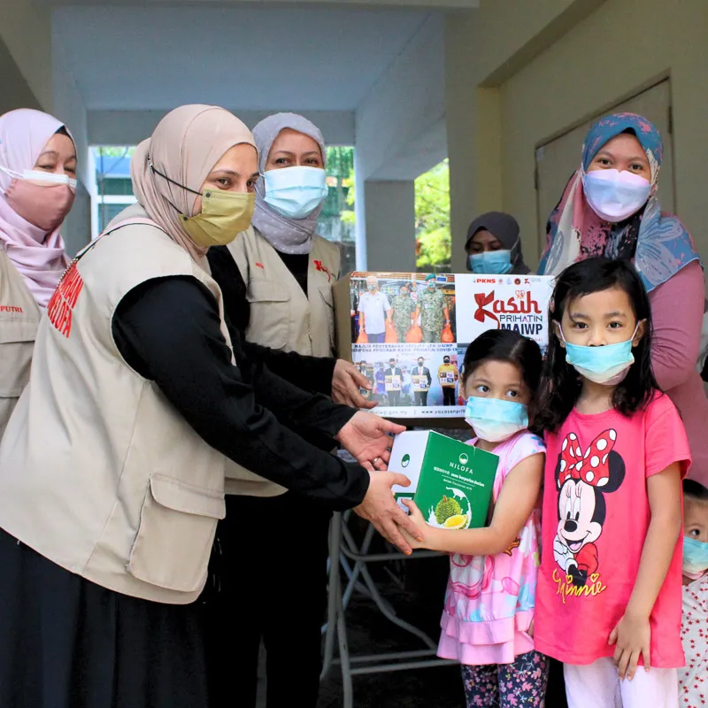 Women wearing hijabs and face masks handing a box to two young girls also wearing face masks indoors.
