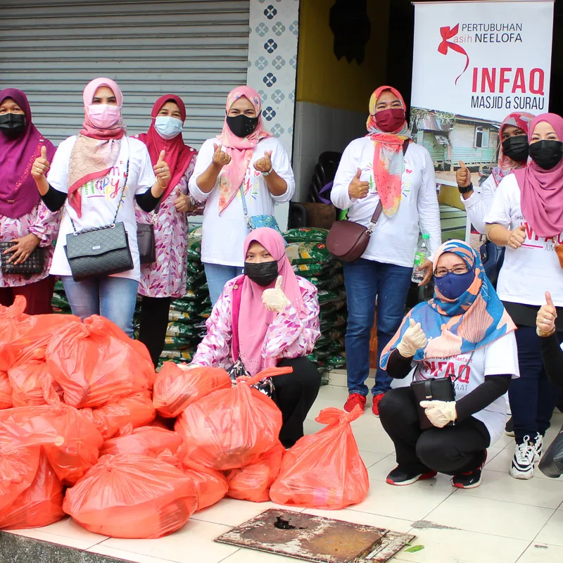 Group of women wearing hijabs and face masks giving thumbs up beside stacked red plastic bags outside a storefront with a banner for Pertubuhan Kasih Neelofa INFAQ Masjid & Surau.