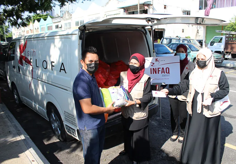 A man receiving a food package from a woman in a red hijab and beige vest in front of a white van with 'Kelofa' written on it, while two other masked women hold signs and envelopes nearby.