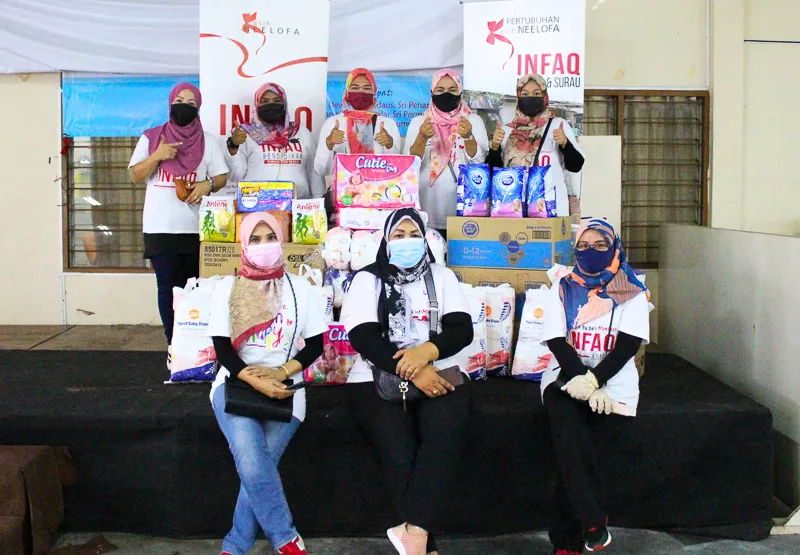 Group of women wearing face masks and hijabs posing with donated food supplies and cardboard boxes in front of banners displaying 'Infaq'.