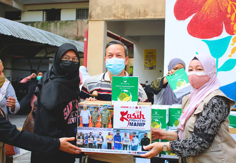 People wearing face masks distributing a box of Nilofa durian drink packs during a community event.