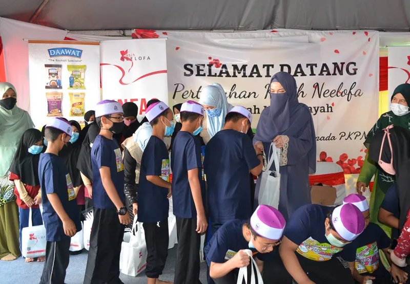 Children wearing purple hats and navy shirts line up to receive gift bags from women in hijabs at an event with a banner saying 'Selamat Datang.'